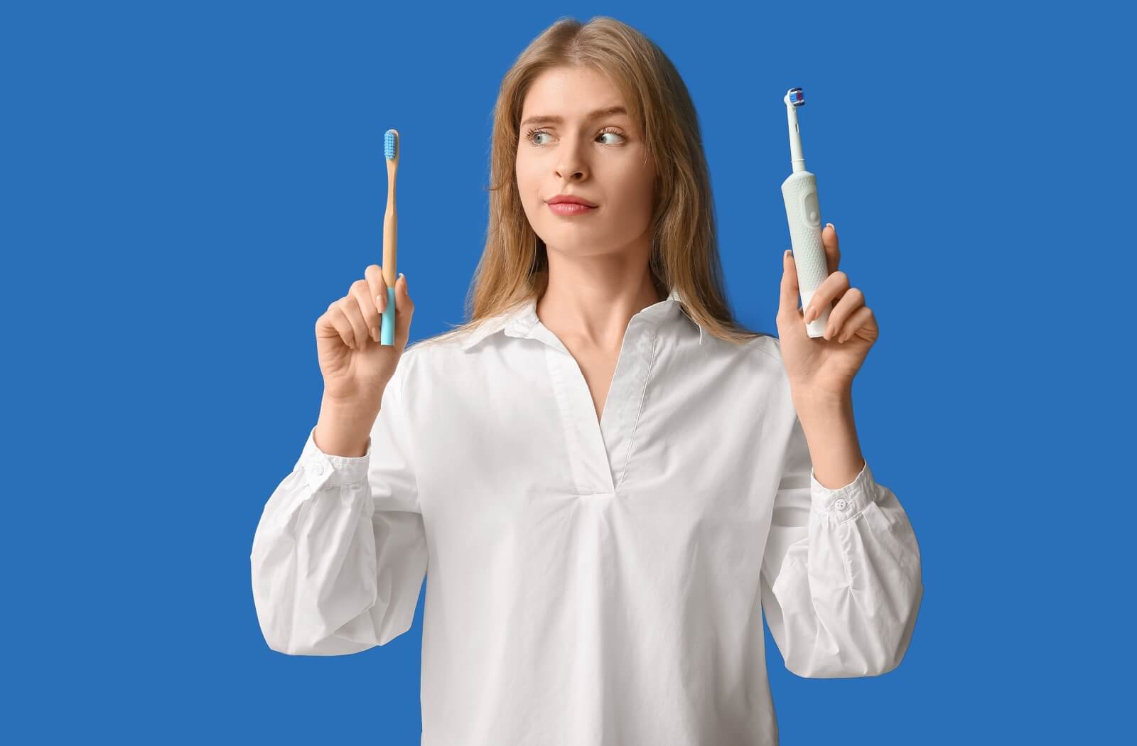 Person holding an electric toothbrush and a manual toothbrush, looking skeptically at the manual toothbrush against a blue background.