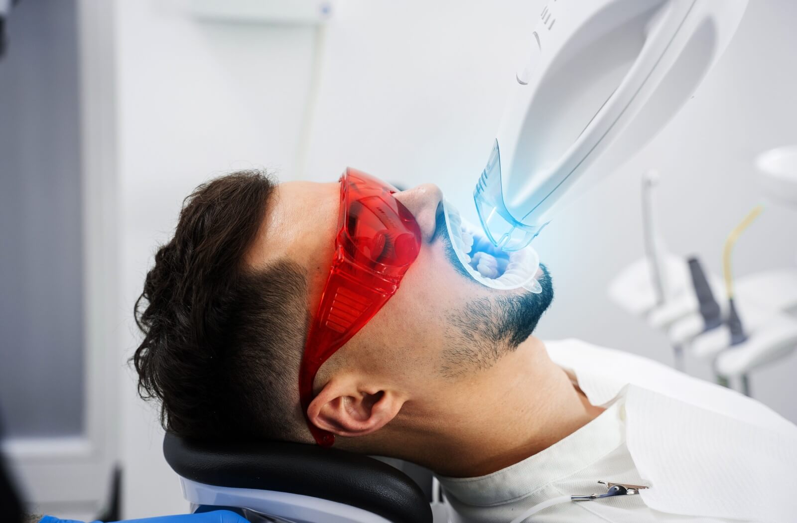 A dentist performing Zoom Whitening on a patient wearing protective red glasses with a bright blue light applied to their teeth