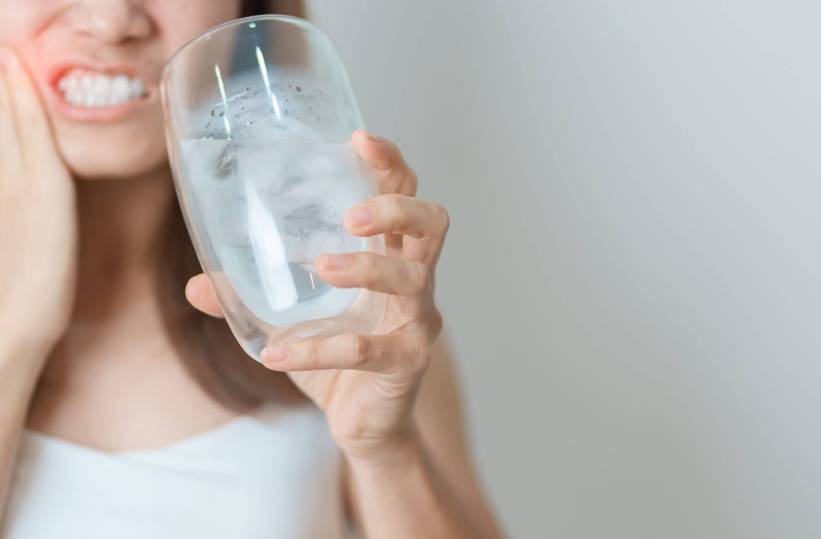 A woman holding a glass of ice water and wincing in pain while touching her cheek, illustrating tooth sensitivity caused by receding gums.