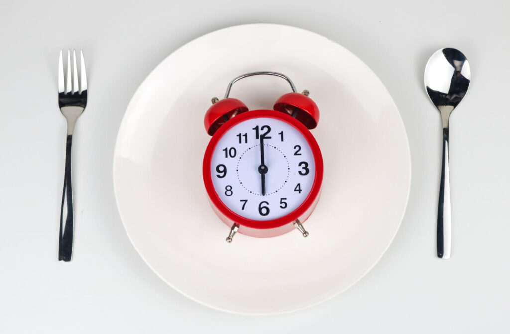 A red alarm clock centered on a white plate between a silver fork and spoon, symbolizing meal timing and recovery schedules.